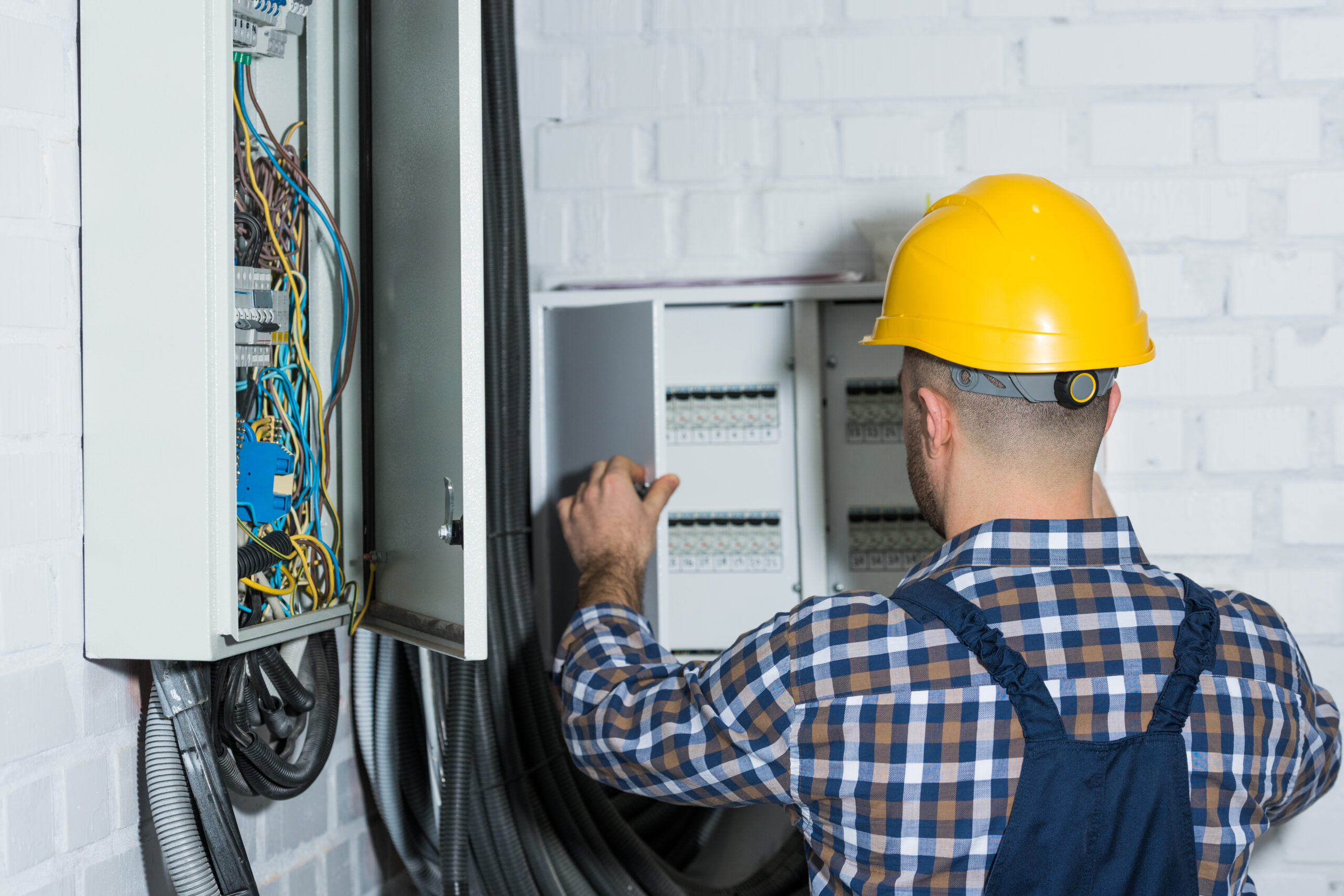 Ein männlicher Elektriker mit einem gelben Helm klemmt eine Leitung im Stromkasten an.