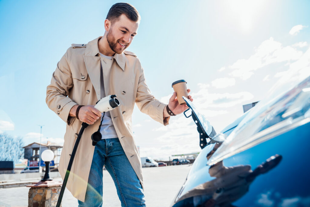 Ein fröhlicher junger Mann mit einem Kaffeebecher in der Hand und einem eleganten Mantel, steckt ein Ladekabel an ein Elektroauto im Sonnenaufgang an.